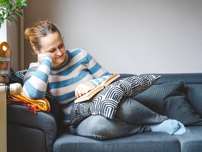 Buch lesende Frau auf Sofa. Sie hat einen Pullover an, es sieht gemütlich aus. 