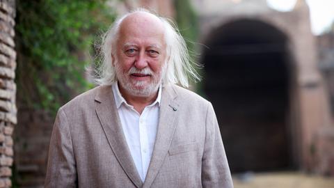 László Krasznahorkai trägt ein beiges Sakko und steht neben einer Mauer im Aussenraum. Er schaut freundlich in die Kamera. László Krasznahorkai trägt ein beiges Sakko und steht neben einer Mauer im Aussenraum. Er schaut freundlich in die Kamera.