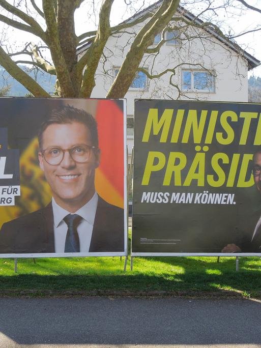 Zwei Wahlplakate der Kandidaten Manuel Hagel und Cem Özdemir vor einem Baum und einem Wohnhaus in Freiburg/Breisgau.