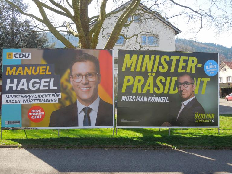 Zwei Wahlplakate der Kandidaten Manuel Hagel und Cem Özdemir vor einem Baum und einem Wohnhaus in Freiburg/Breisgau.