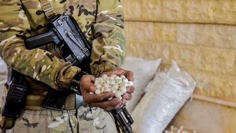 Ein Soldat mit einem Maschinengewehr quer vor dem Körper hängend hält beide Hände voll mit weißen Pillen in die Kamera. Im Hintergrund stehen weiße Säcke, aus denen weiße Pillen rollen. Ein Soldat mit einem Maschinengewehr quer vor dem Körper hängend hält beide Hände voll mit weißen Pillen in die Kamera. Im Hintergrund stehen weiße Säcke, aus denen weiße Pillen rollen.