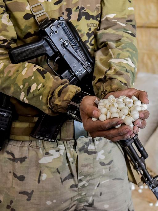 Ein Soldat mit einem Maschinengewehr quer vor dem Körper hängend hält beide Hände voll mit weißen Pillen in die Kamera. Im Hintergrund stehen weiße Säcke, aus denen weiße Pillen rollen.