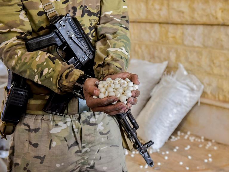 Ein Soldat mit einem Maschinengewehr quer vor dem Körper hängend hält beide Hände voll mit weißen Pillen in die Kamera. Im Hintergrund stehen weiße Säcke, aus denen weiße Pillen rollen.
