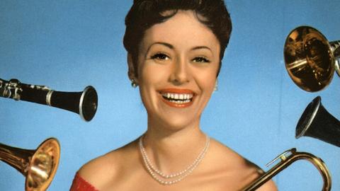 Caterina Valente steht im roten Kleid lachend vor einer blauen Wand und wird von allerlei Blasinstrumenten umflort