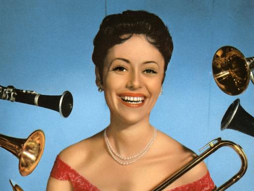 Caterina Valente steht im roten Kleid lachend vor einer blauen Wand und wird von allerlei Blasinstrumenten umflort