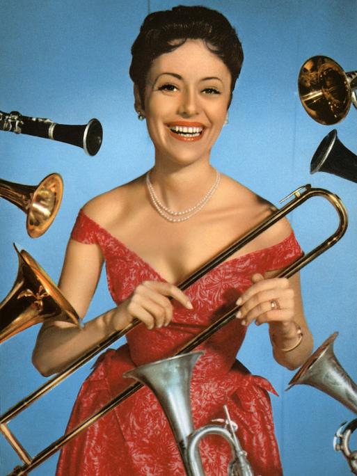 Caterina Valente steht im roten Kleid lachend vor einer blauen Wand und wird von allerlei Blasinstrumenten umflort