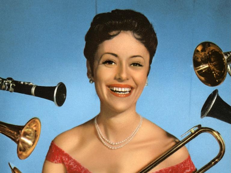 Caterina Valente steht im roten Kleid lachend vor einer blauen Wand und wird von allerlei Blasinstrumenten umflort