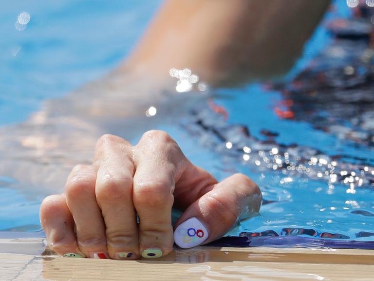 Eine Fünfkampf-Athletin fasst mit ihrer Hand an den Beckenrand. Ihre Nägel sind im Stil der Olympischen Ringe lackiert. 