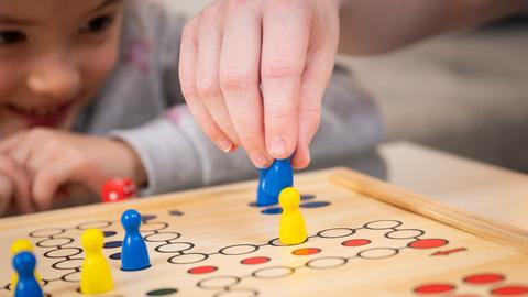 Nahaufnahme: Eine Kinderhand hält eine Spielfigur und schlägt damit eine andere aus dem Spiel "Mensch ärgere dich nicht" heraus, im Hintergrund sieht ein Mädchen gespannt zu und lacht.