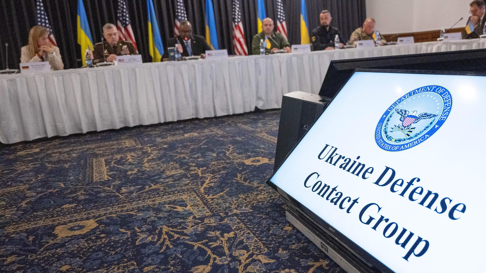 Ein Bildschirm mit der Aufschrift "Ukraine Defense Contact Group", dahinter ist ein Tisch mit mehreren Menschen vor verschiedenen Flaggen zu sehen. 