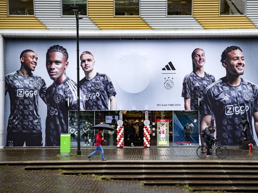 Blick von Außen auf einen Ajax-Fanshop in der Johan Cruijff ArenA in Amsterdam. An der Fassade ist ein großes Poster von Ajax-Spielern zu sehen. 