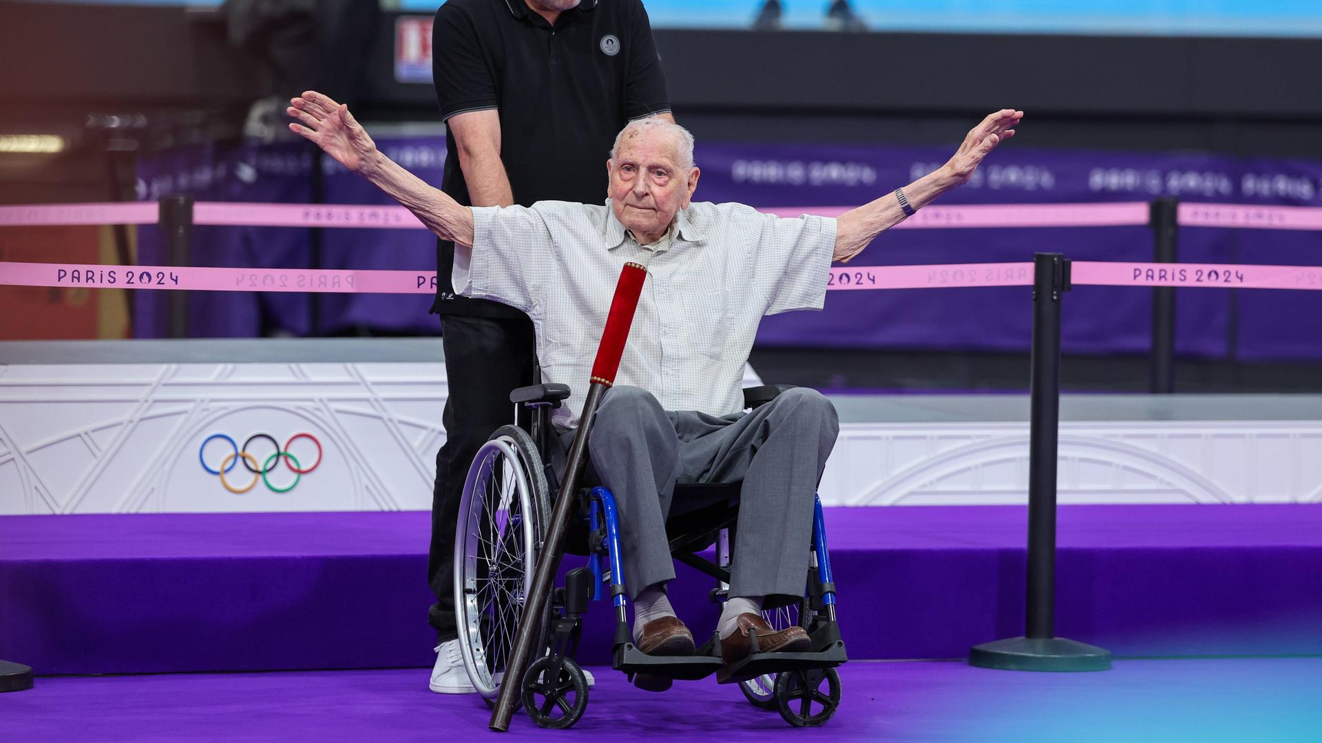 Chales Coste sitzt in einem Rollstuhl und hebt grüßend die Arme. Chales Coste sitzt in einem Rollstuhl und hebt grüßend die Arme.