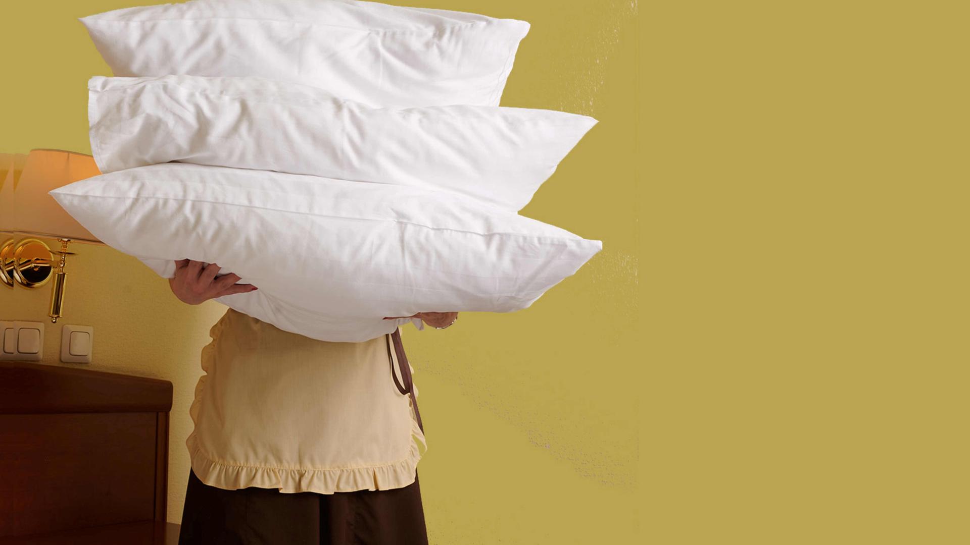 Eine Person mit Schürze hält drei Kopfkissen vor ihren Körper. Eine Person mit Schürze hält drei Kopfkissen vor ihren Körper.