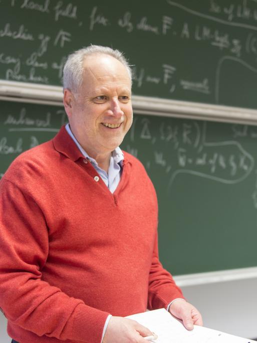 Michale Rapoport steht vor einer Tafel, die mit Formeln beschrieben ist, trägt einen roten Pullover, hält Papier und Stift in der Hand und schaut freundlich an der Kamera vorbei.