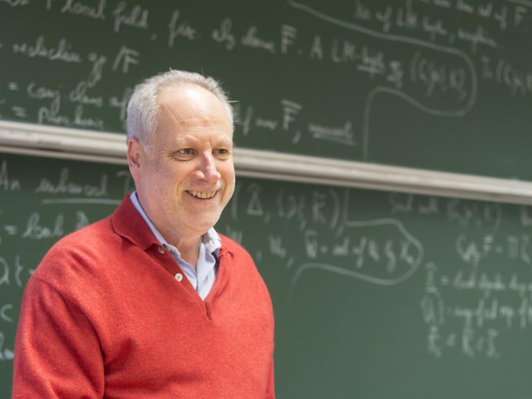 Michale Rapoport steht vor einer Tafel, die mit Formeln beschrieben ist, trägt einen roten Pullover, hält Papier und Stift in der Hand und schaut freundlich an der Kamera vorbei.