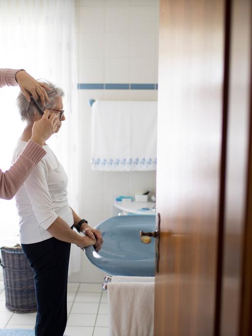 Eine alte Frau steht in einem Badezimmer am Waschbecken. Eine junge Frau steht hinter der Seniorin und kämmt ihr das Haar. Eine alte Frau steht in einem Badezimmer am Waschbecken. Eine junge Frau steht hinter der Seniorin und kämmt ihr das Haar.