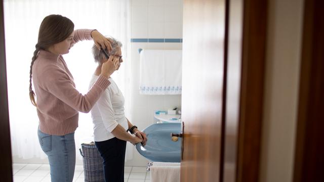 Eine alte Frau steht in einem Badezimmer am Waschbecken. Eine junge Frau steht hinter der Seniorin und kämmt ihr das Haar. Eine alte Frau steht in einem Badezimmer am Waschbecken. Eine junge Frau steht hinter der Seniorin und kämmt ihr das Haar.