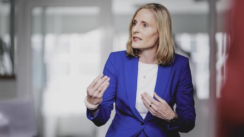 Das Bild zeigt Marie-Christine Ostermann während eines Interviews auf einem Stuhl sitzend in einem Raum. Sie trägt einen blauen Business-Anzug und unterstreicht ihre Aussagen, indem sie mit beiden Händen gestikuliert
