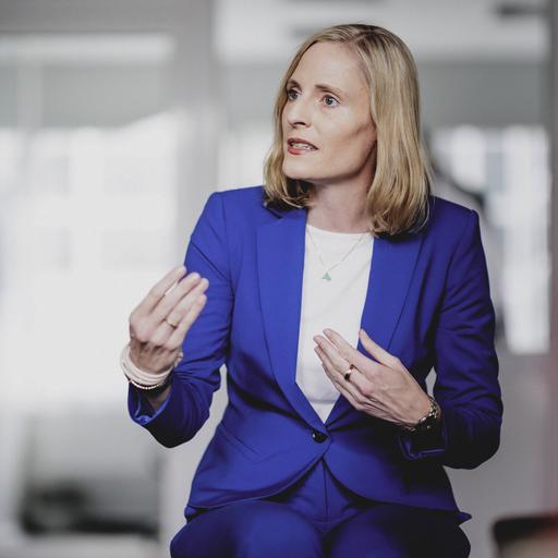 Das Bild zeigt Marie-Christine Ostermann während eines Interviews auf einem Stuhl sitzend in einem Raum. Sie trägt einen blauen Business-Anzug und unterstreicht ihre Aussagen, indem sie mit beiden Händen gestikuliert