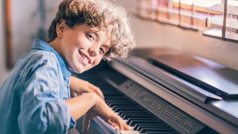 Eine Junge mit braunen Locken lacht in die Kamera, während er auf einem elektrischen Klavier spielt.
