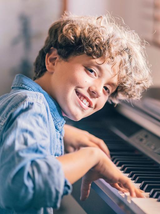 Eine Junge mit braunen Locken lacht in die Kamera, während er auf einem elektrischen Klavier spielt.