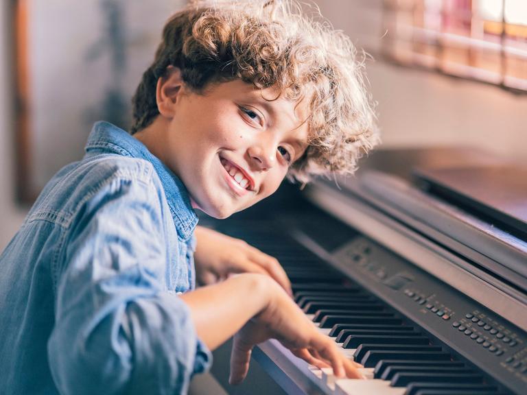 Eine Junge mit braunen Locken lacht in die Kamera, während er auf einem elektrischen Klavier spielt.