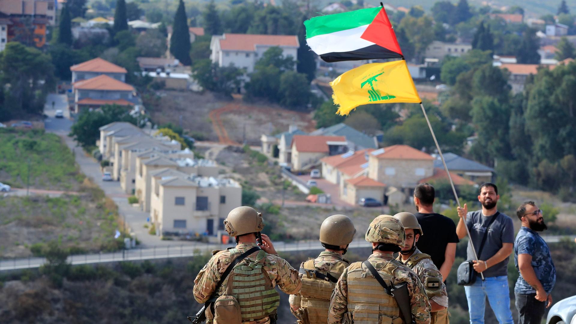 Libanon, Kafr Kila: Libanesische Soldaten stehen auf einem Hügel mit Blick auf die israelische Siedlung Mutelh, während ein Mann die palästinensische und die Hisbollah-Flagge in der Nähe der libanesisch-israelischen Grenze schwenkt. 