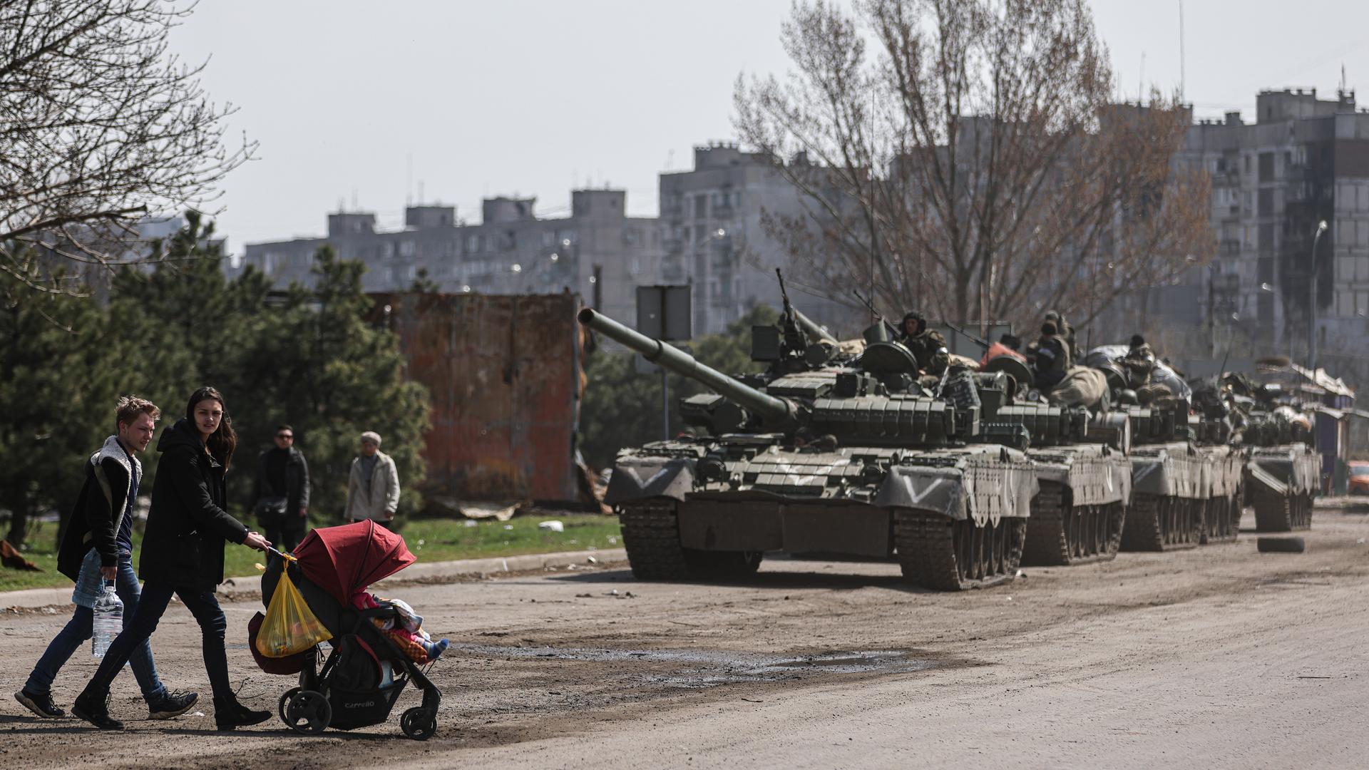 Mariupol im April 2022: Zivilisten mit Kinderwagen überqueren die Straße vor mehreren russischen Panzern