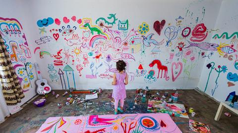 Ein junges Mädchen steht in einem Zimmer vor einer über und über mit bunten Motiven bemalten Wand.