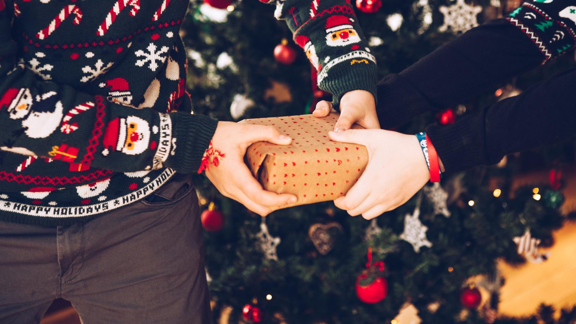 Zwei Personen mit weihnachtlichen Pullis ziehen vor einem Weihnachtsbaum an einem Geschenk.