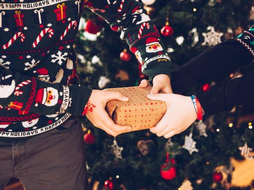 Zwei Personen mit weihnachtlichen Pullis ziehen vor einem Weihnachtsbaum an einem Geschenk.