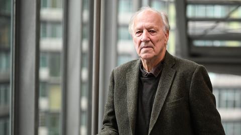 Werner Herzog steht mit ernster Miene vor einer Glasfassade an einem Treppengeländer