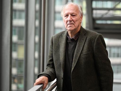 Werner Herzog steht mit ernster Miene vor einer Glasfassade an einem Treppengeländer