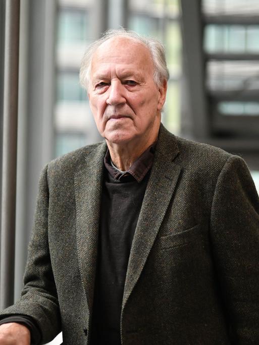 Werner Herzog steht mit ernster Miene vor einer Glasfassade an einem Treppengeländer