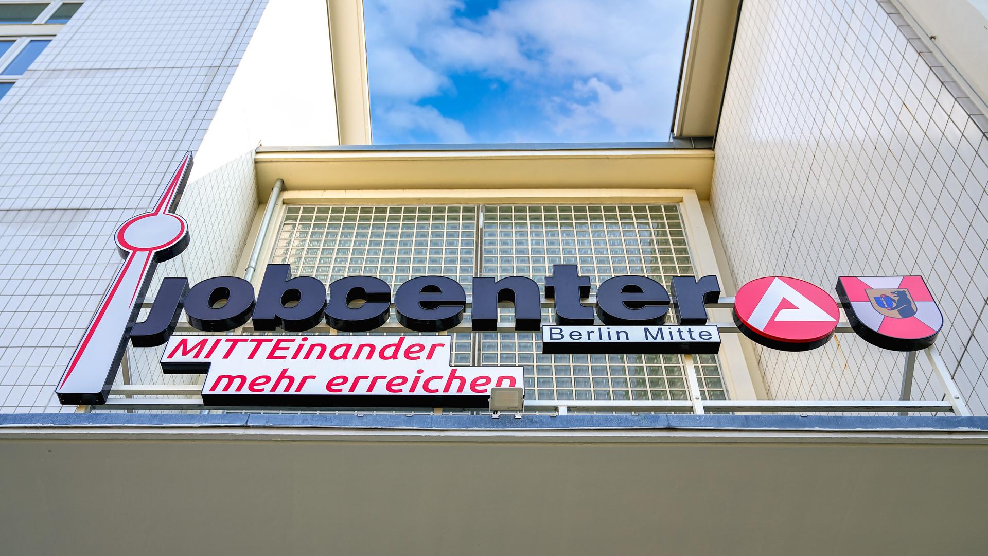 Eingangsbereich vom Jobcenter mit dem Jobcenter-Logo dem Logo der Agentur für Arbeit, einem Berliner Wappen, dem Slogan "MITTEinander mehr erreichen" und den markanten Glasbausteinen in der Fassade. 