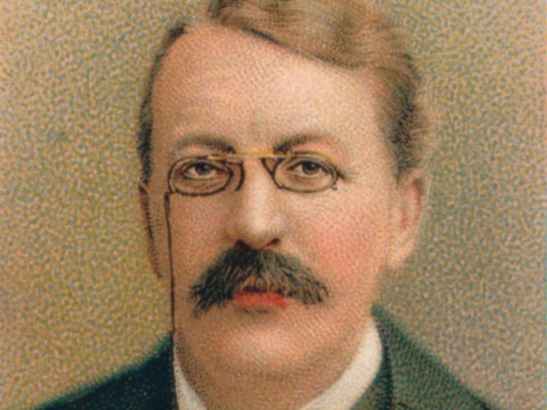 Nochkoloriertes Porträt von  Sir Charles Villiers Stanford, das wohl um 1911 entstand und ihn mit Brille und vornehmen Anzug zeigt.
