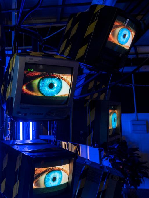 Eine Installation von acht Fernsehbildschirmen, die in einem Halbkreis angeordnet sind, zeigen jeweils die gleiche Nahaufnahme eines menschlichen Auges mit einer blauen Iris.