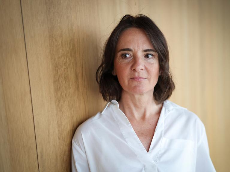 Martina Heßler mit weißer Bluse und dunklen Haaren steht vor einer Wand aus Holz und schaut zur Seite