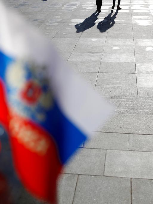 Eine unscharf fotografierte russische Flagge flattert im Wind. Am oberen Bildrand sihet man lange Schlagschatten von Personen.
