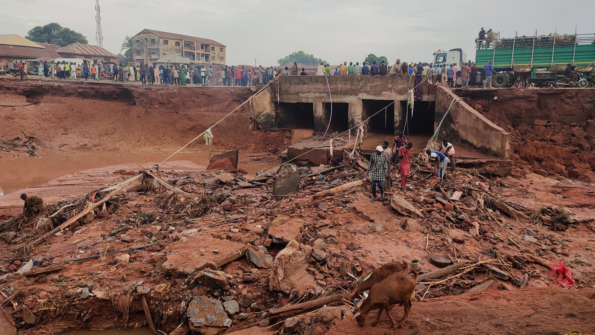 Menschen stehen in einem überfluteten Gebiet in Mokwa, Nigeria auf rötlichem Schlamm und Geröll. Dahinter eine abgerutschte Straße, auf der zahlreiche weitere Menschen stehen. 
