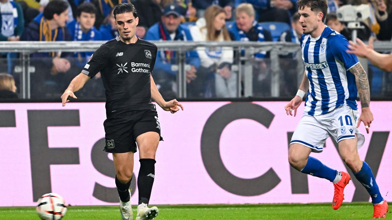 Patrik Walemark von Lech Posen und Miguel Gutierrez von Bayer 04 Leverkusen rennen nebeneinander dem Ball hinterher. Dahinter Zuschauer auf einer Tribüne. 
