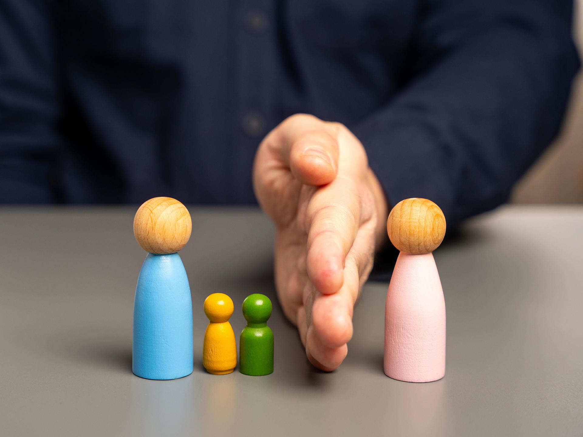 Eine Hand trennt eine blau angemalte Holzfigur zusammen mit zwei kleineren Holzfiguren von einer rosa Holzfigur.