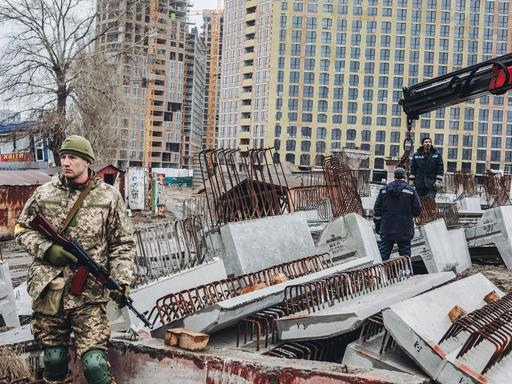 Der Soldat in Tarnanzug steht vor den Betonteilen. Eines davon wird gerade von zwei Arbeitern mit einem Kran heruntergelassen. Im Hintergrund Hochhäuser.