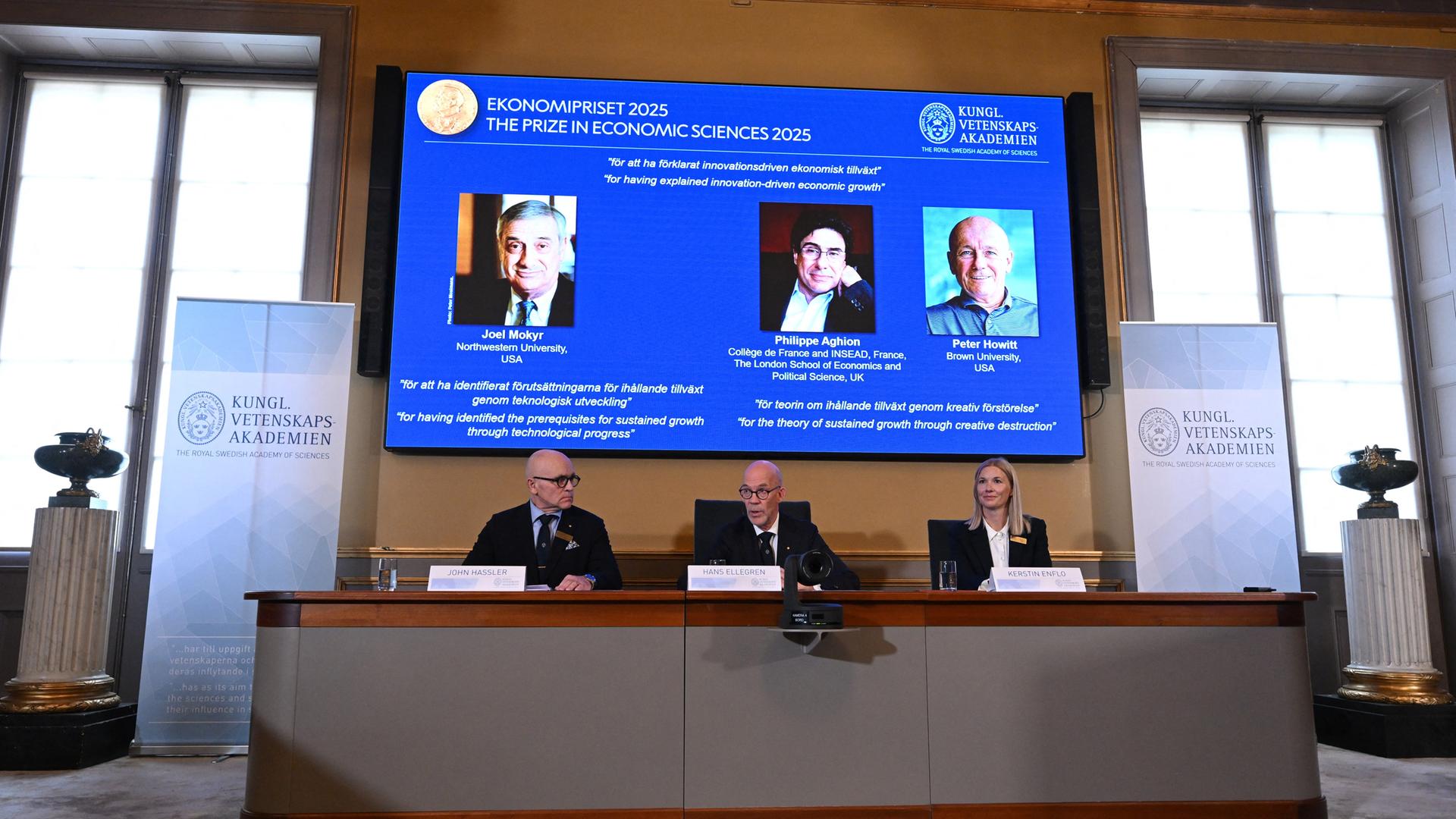 Die drei Preisträger sind auf einer blauen Leinwand zu sehen. Darunter sitzen drei Mitglieder der Schwedischen Akademie.
