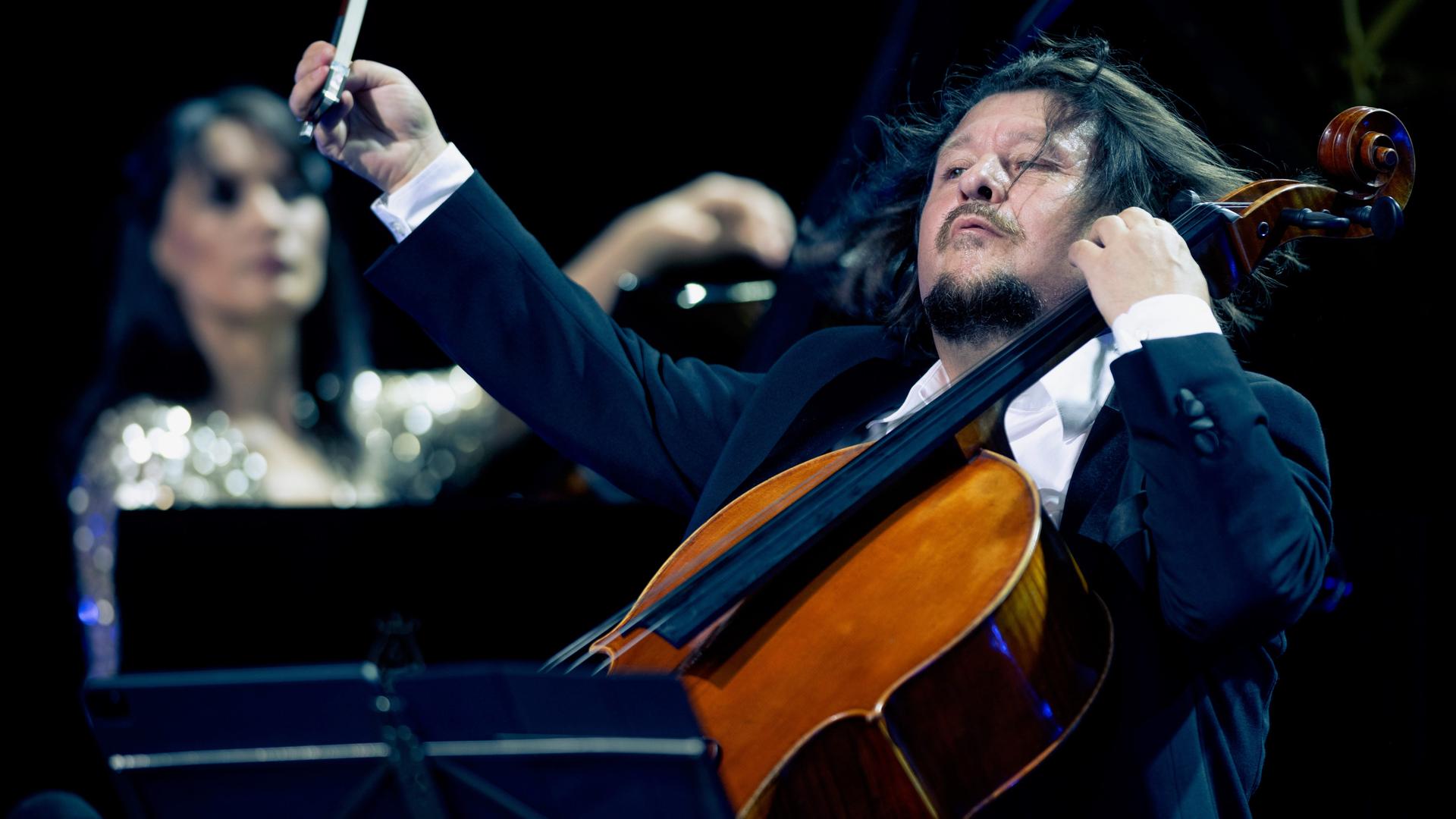 Im Vordergrund spielt Friedrich Kleinhapl mit einer wilden Geste auf seinem Violoncello. Im Hintergrund ist verschwommen Kima Barbier am Klavier zu erkennen. Im Vordergrund spielt Friedrich Kleinhapl mit einer wilden Geste auf seinem Violoncello. Im Hintergrund ist verschwommen Kima Barbier am Klavier zu erkennen.