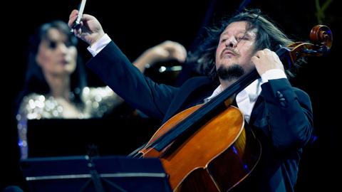 Im Vordergrund spielt Friedrich Kleinhapl mit einer wilden Geste auf seinem Violoncello. Im Hintergrund ist verschwommen Kima Barbier am Klavier zu erkennen.