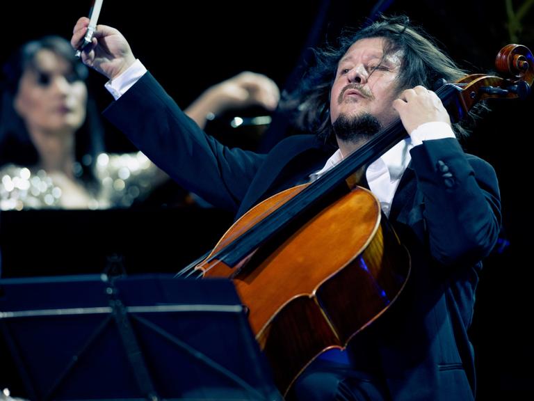 Im Vordergrund spielt Friedrich Kleinhapl mit einer wilden Geste auf seinem Violoncello. Im Hintergrund ist verschwommen Kima Barbier am Klavier zu erkennen.