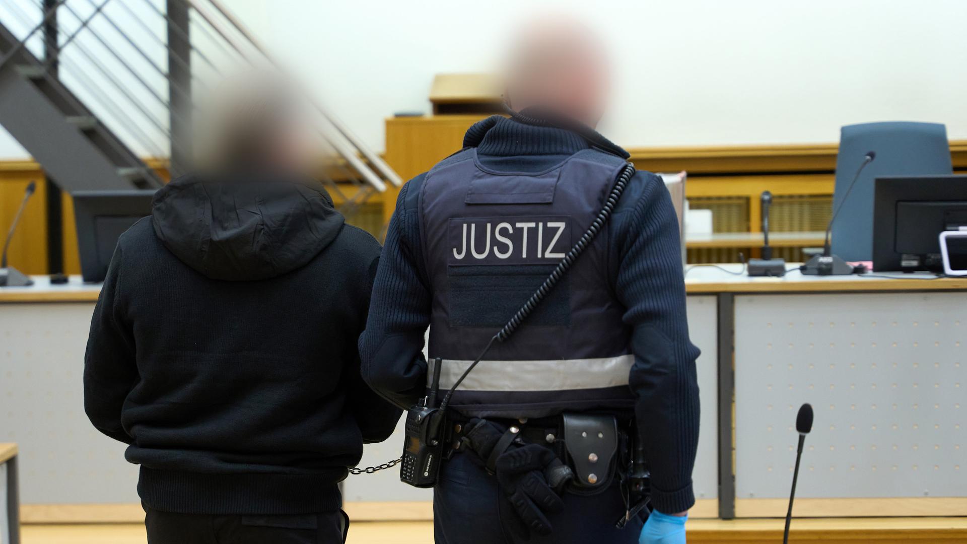 Ein Justizvollzugsbeamter führt einen Mann, dem Beihilfe zum Mord und Beihilfe zu versuchtem Mord vorgeworfen wird, in den Gerichtssaal des Oberlandesgerichts Koblenz.