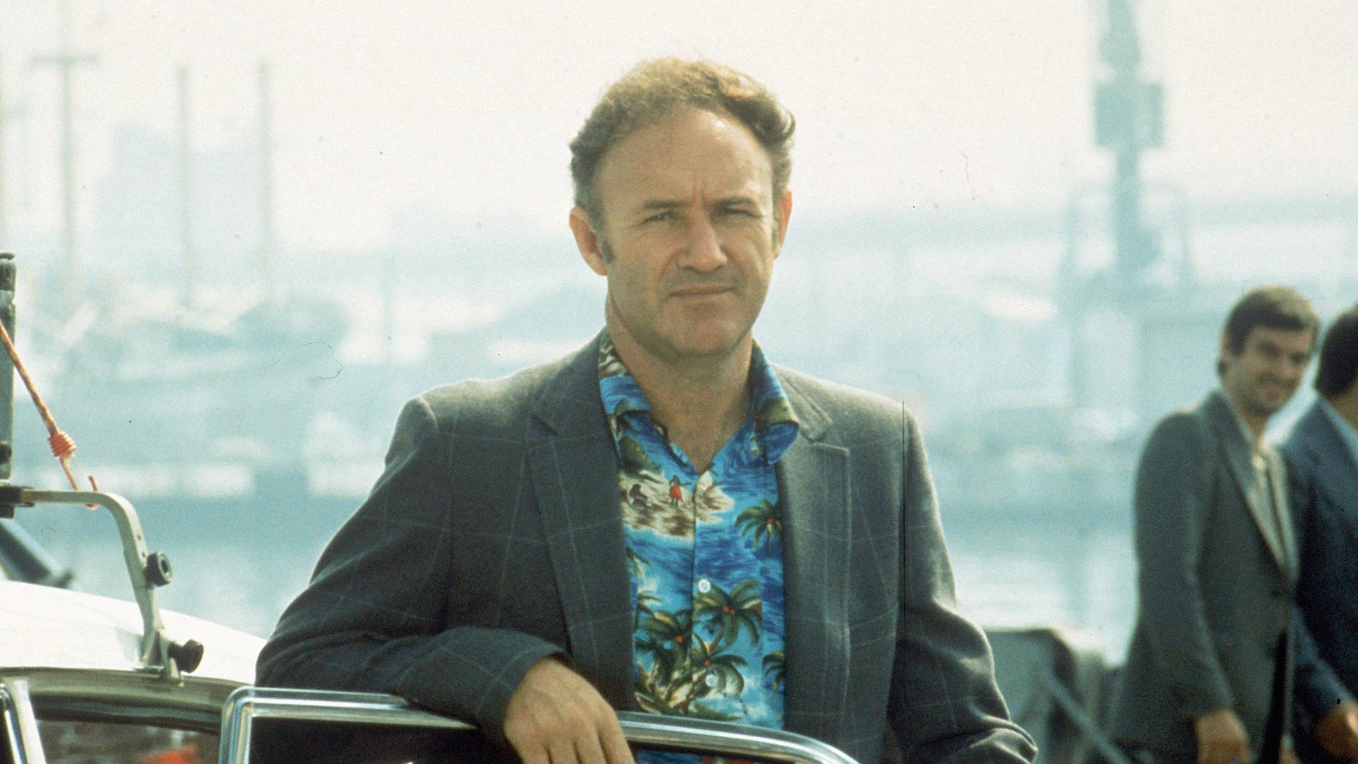 Aufnahme aus dem Film „The French Connection“. Es zeig den Schauspieler Gene Hackman in Hawaii-Hemd und Sakko an eine offene Autotür gelehnt.