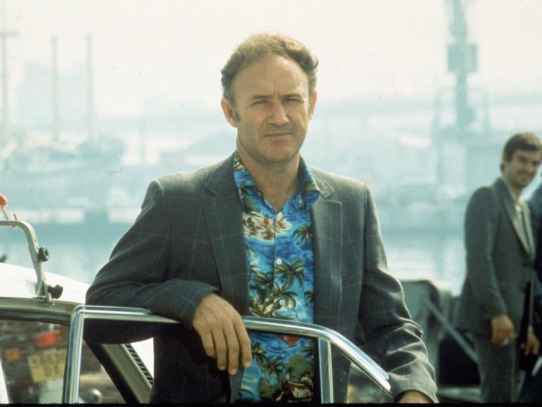 Aufnahme aus dem Film „The French Connection“. Es zeig den Schauspieler Gene Hackman in Hawaii-Hemd und Sakko an eine offene Autotür gelehnt.